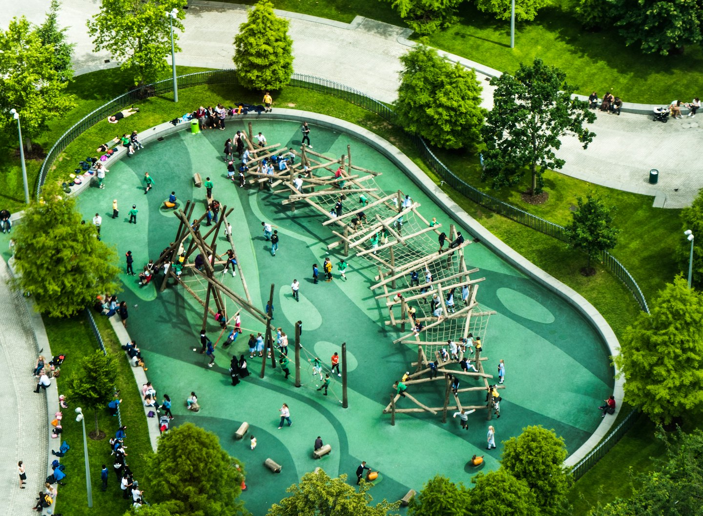 Children and adults play on a modern wooden playground on a sunny day.
